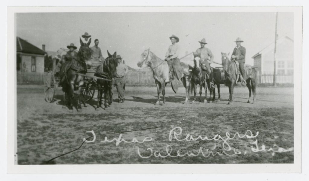 Black and white photo of seven men. On the left three sit in a horse-drawn wagon while one leans on the wagon’s side. On the right three are on horseback. A cursive caption in white at the bottom of the photo reads, “Texas Rangers Valentine, Texas.”