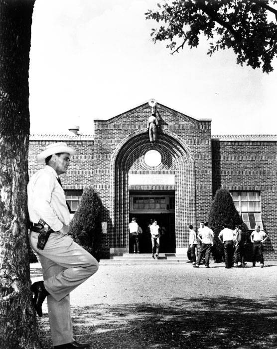 Black-and-white photo of Banks outside of Mansfield High School while the effigy hangs over the main entrance. Banks leans against a tree with one foot resting on the trunk and looks relaxed.