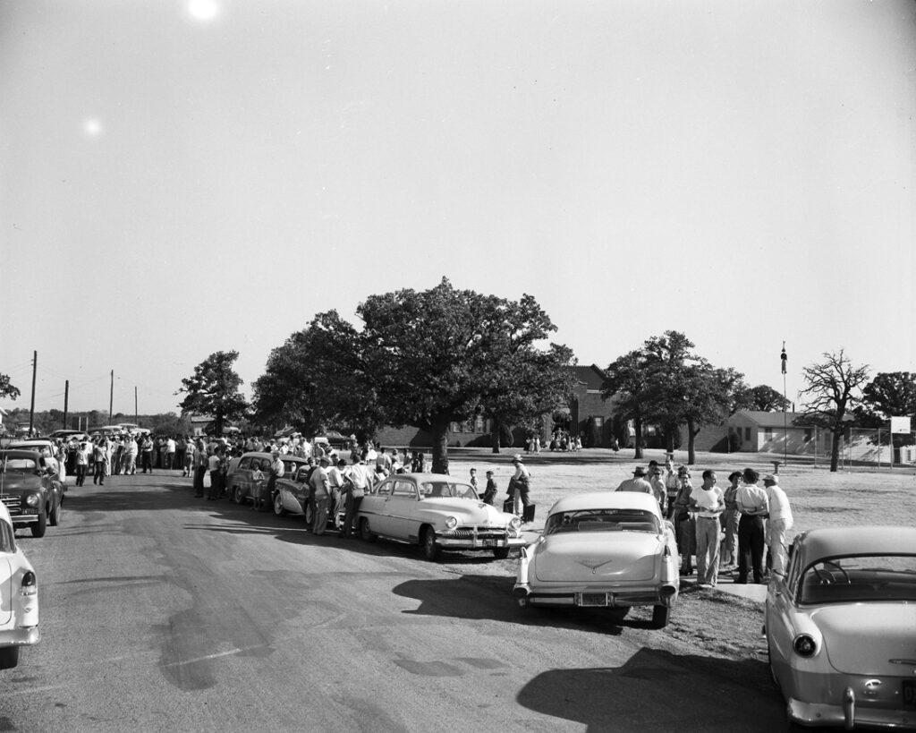 Black-and-white photo of 1950s-era cars parked on either side of a road with crowds of white people. To the right of the road is a yard with school buildings. The effigy of a Black person hangs from a flagpole in the yard. From The Crisis at Mansfield, University of North Texas.