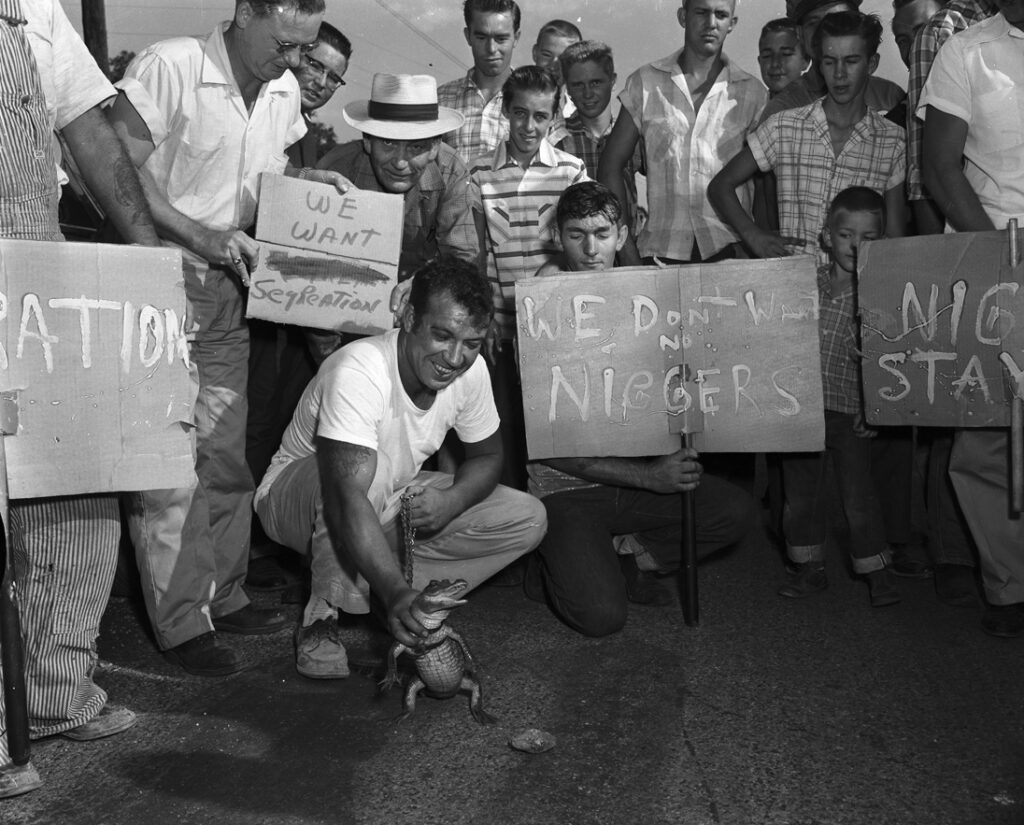 Black-and-white photo of white men and boys gathered around a white man holding a baby alligator. The people in the crowd hold signs reading “we want segregation” and “we don’t want no [n-word].”