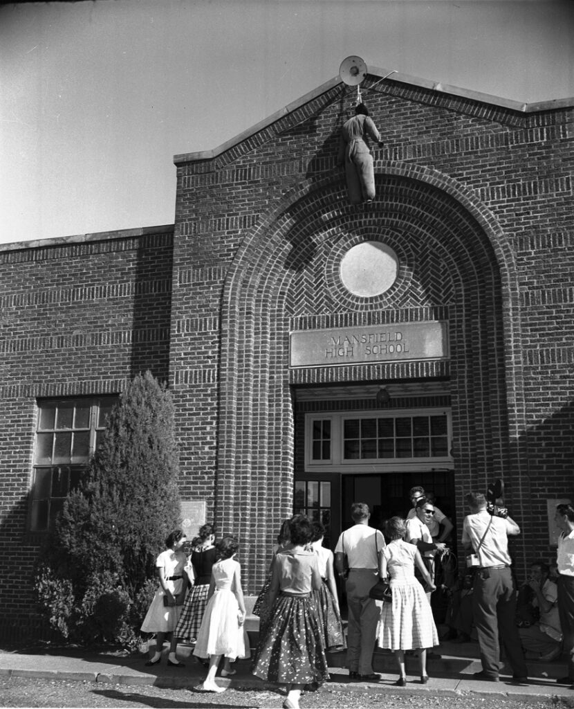 Black-and-white photograph of white students in 1950-style clothes entering Mansfield High School, a one-story brick building with an arched entrance. An effigy of a Black person is hung from the bullhorn at the top of the arch.