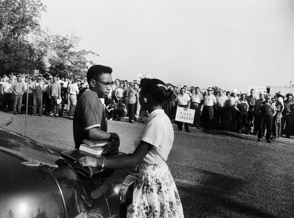 Black-and-white photo of two Black teenagers, a boy and a girl, standing in the foreground at the hood of a car. In front of them across a strip of grass is a large crowd of white adults, mostly men. One holds a sign reading, “No Mixed Classes.”