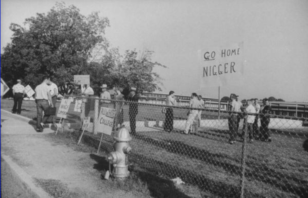 Black-and-white photo of a group of white people, mostly men, milling about on a sidewalk and field. Some hold signs. The only readable one says, “Go Home [N-word].”